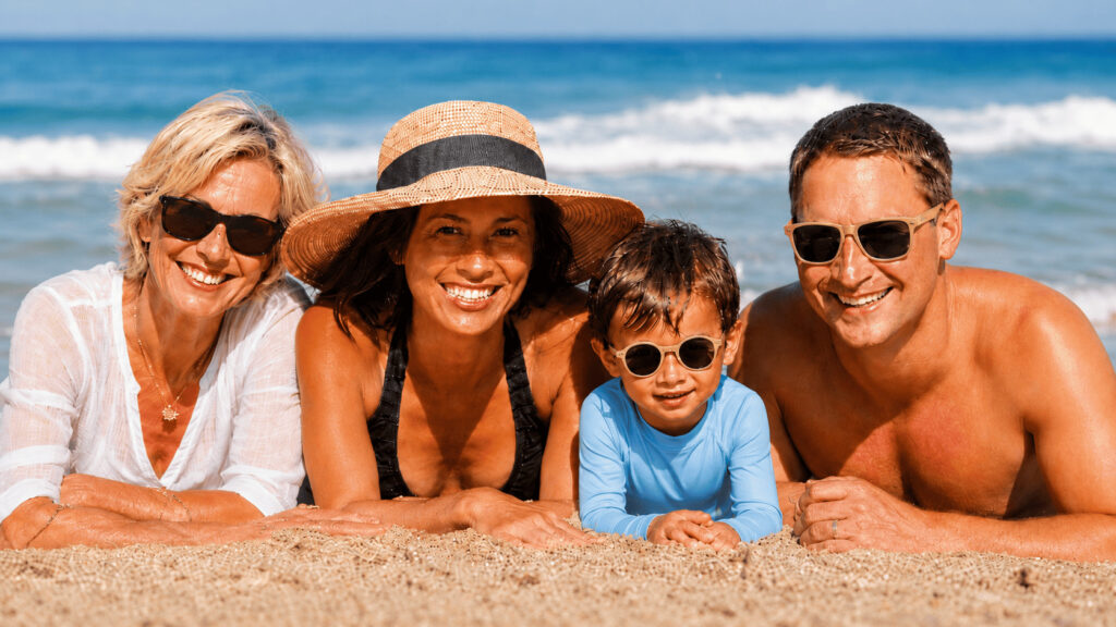 Famille de trois générations allongée sur le sable à la plage, portant lunettes de soleil, chapeau et vêtement anti-UV pour illustrer la protection solaire à tout âge.