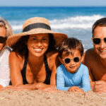 Famille de trois générations allongée sur le sable à la plage, portant lunettes de soleil, chapeau et vêtement anti-UV pour illustrer la protection solaire à tout âge.