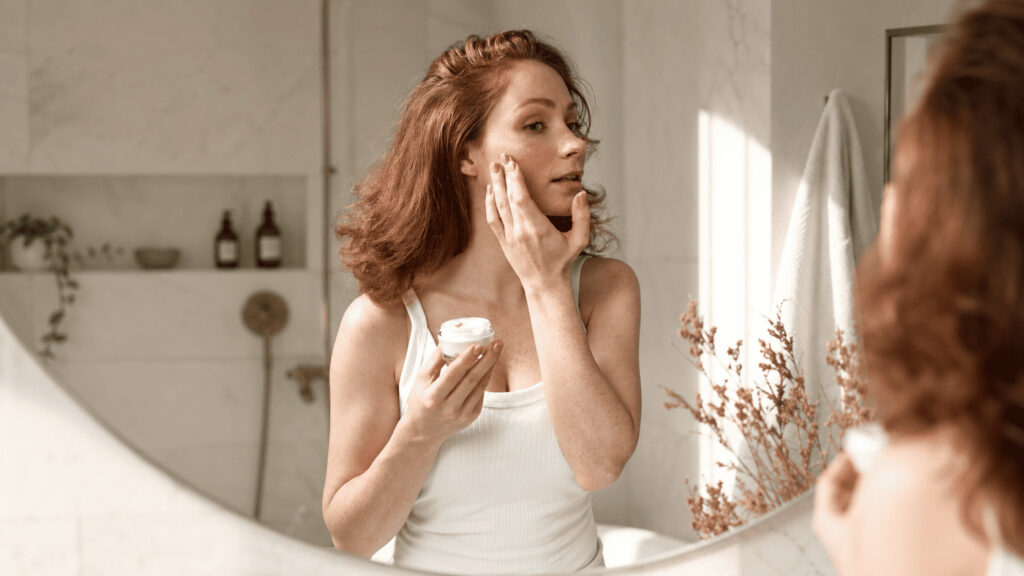 Femme appliquant une crème sur son visage devant le miroir dans une salle de bain lumineuse et minimaliste.
